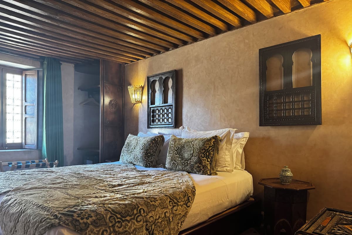 A bedroom with a large bed, patterned bedding, a wooden ceiling, and a wall-mounted light fixture.