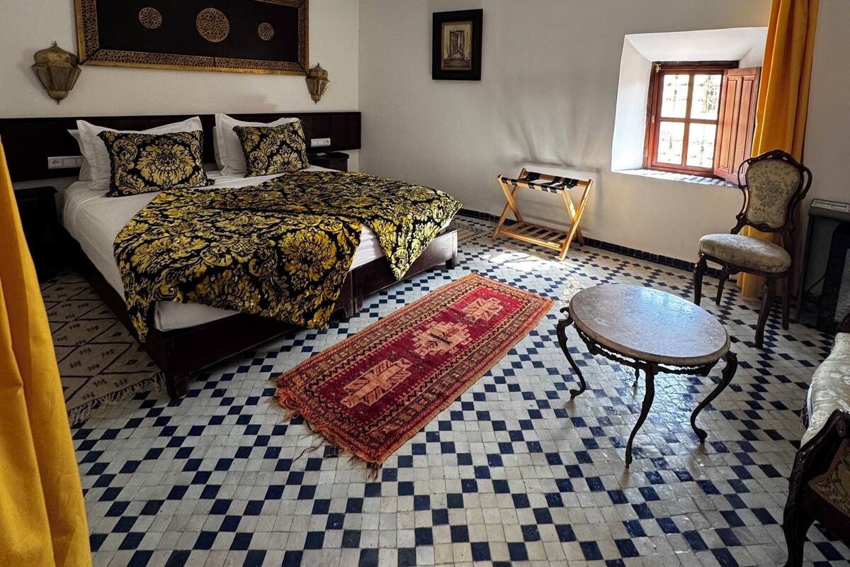 A room with a checkered floor, a bed with patterned bedding, a red rug, a small table, and a chair.
