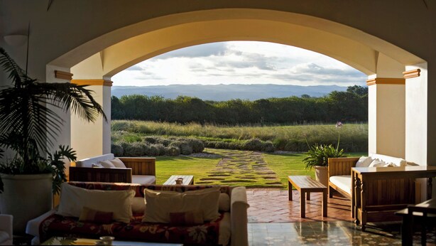 Habitación con vista a un paisaje verde a través de un arco. Se observan varios muebles, como un sofá, una silla y una mesa.