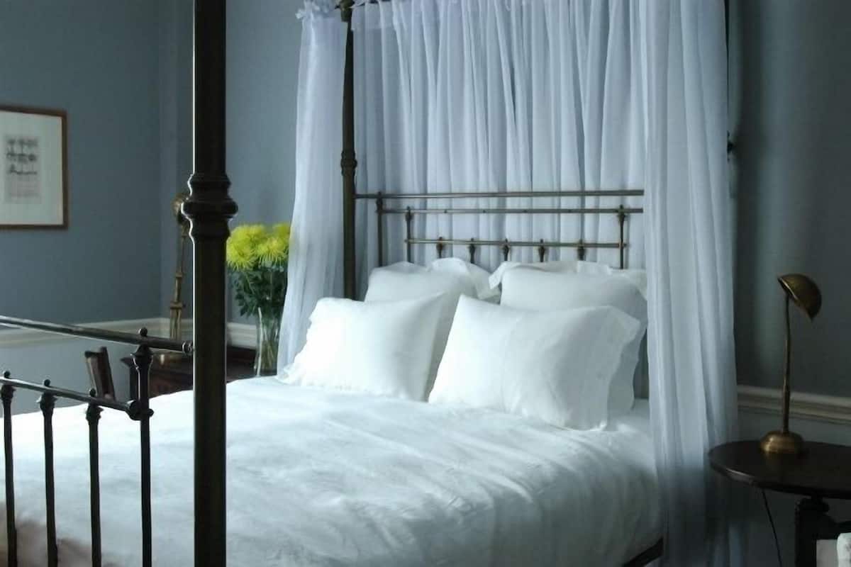 Un lit à baldaquin à quatre poteaux, avec du linge de lit blanc, une tête de lit en métal et une table de chevet avec une lampe.