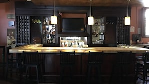 Zona de bar amb una barra llarga, diversos tamborets i llums penjants. Darrere de la barra, hi ha estantes amb ampolles i vasos.