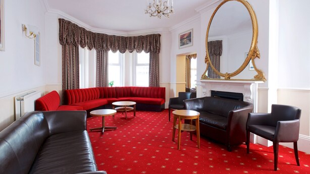 A room with a red carpet, a black leather sofa, a red sectional sofa, a black armchair, and a round wooden table. - Glendevon Hotel (Bournemouth)