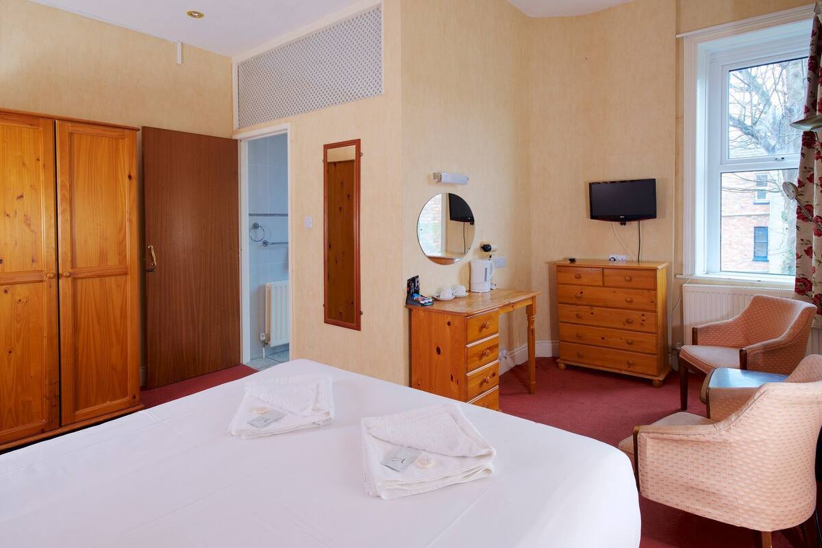 A hotel room with a large bed, wooden wardrobe, a dresser, a television, and a window with a view of buildings.