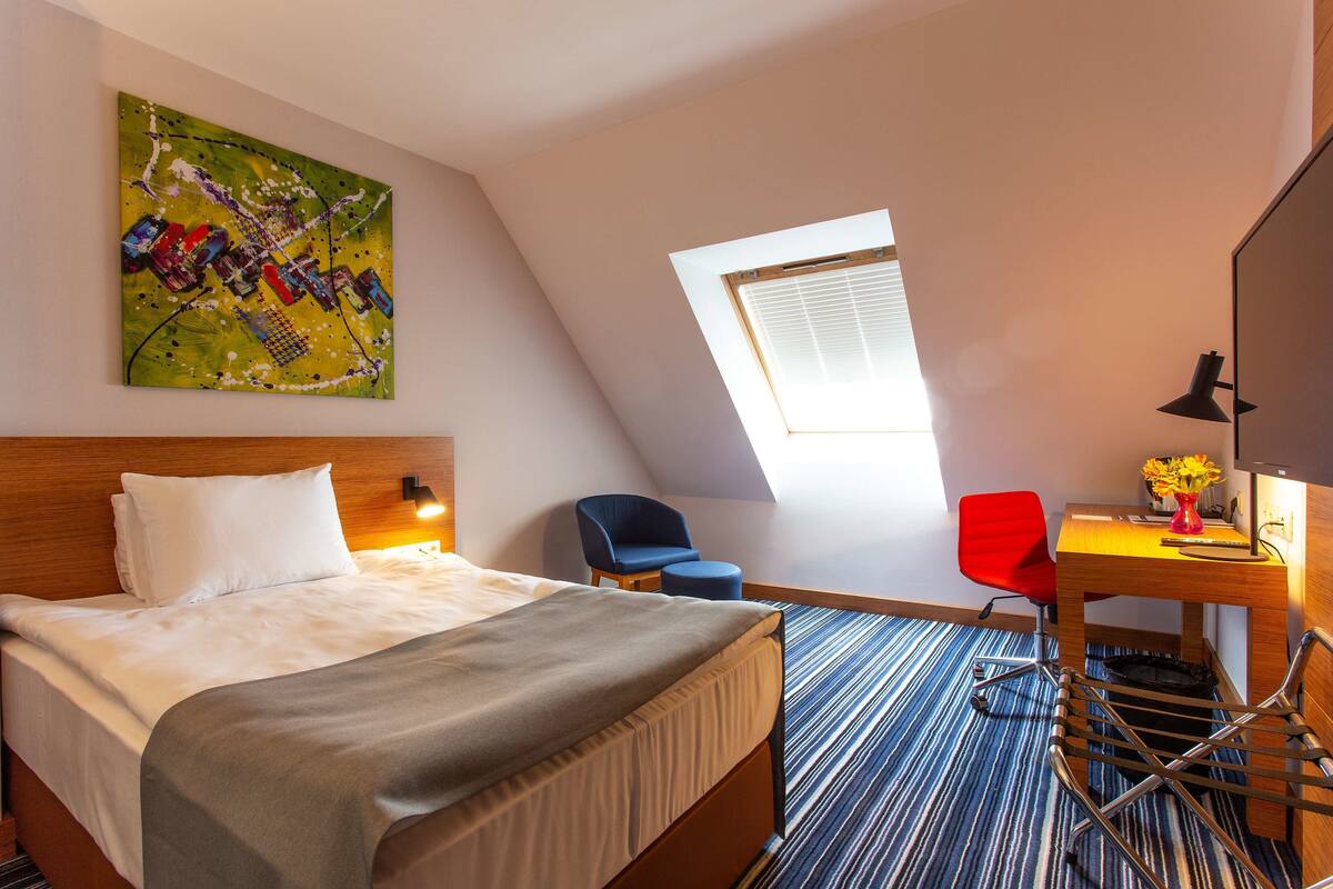 A hotel room with a bed, a desk with a chair, a painting on the wall, and a window.