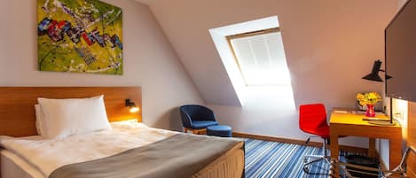 A hotel room with a bed, a desk with a chair, a painting on the wall, and a window.