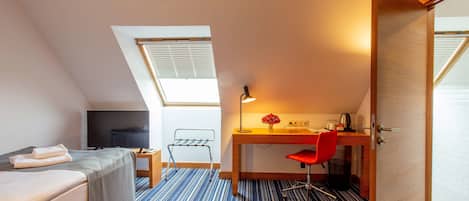 A compact hotel room with a bed, a desk, a chair, a TV, and a small table with a vase.