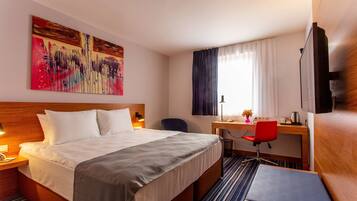 A hotel room with a bed, a desk, a chair, a TV, and a large abstract painting on the wall.
