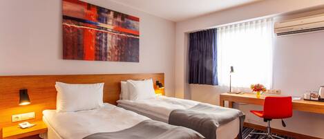 A hotel room with two beds, a desk, a chair, a window with curtains, an air conditioner, and a painting on the wall.