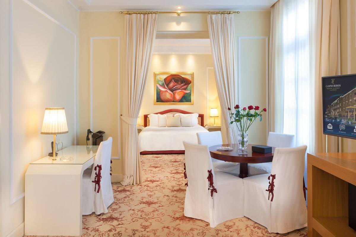A hotel room with a bed, a desk with a lamp, a chair, a table with a vase of flowers, and a painting on the wall.