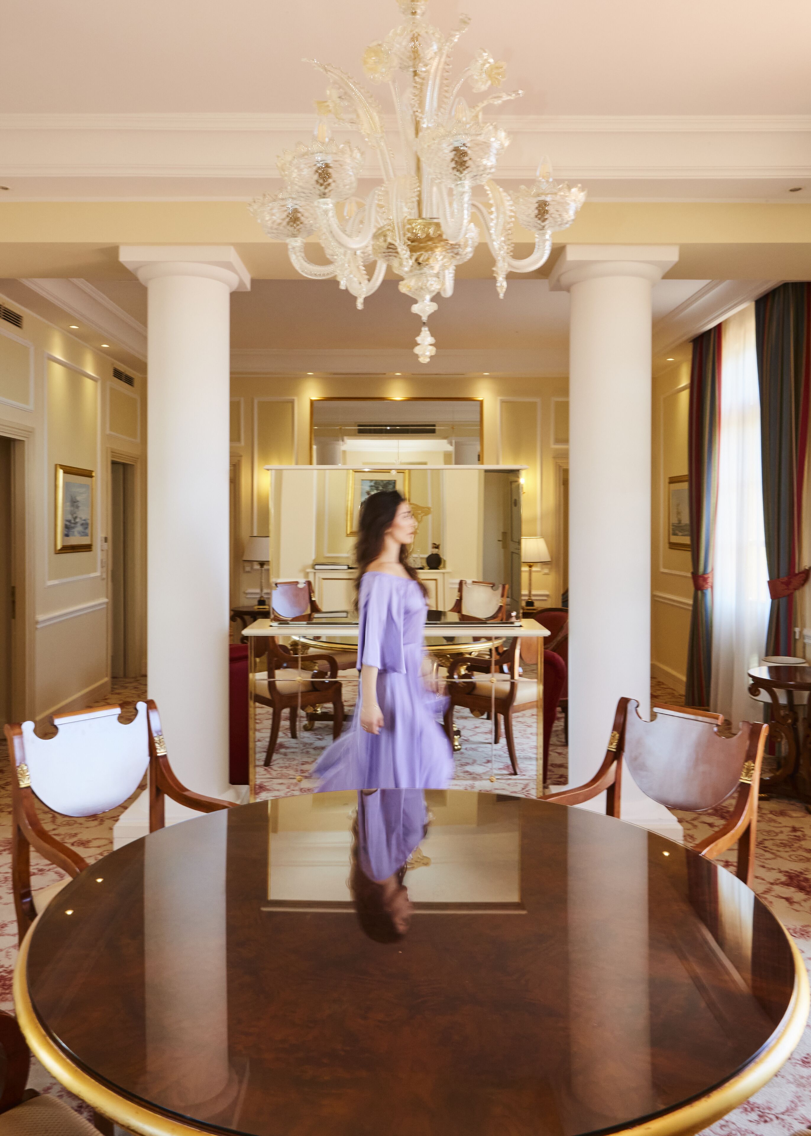 A woman walking through a room with a large round table, chairs, a chandelier, and a mirror.