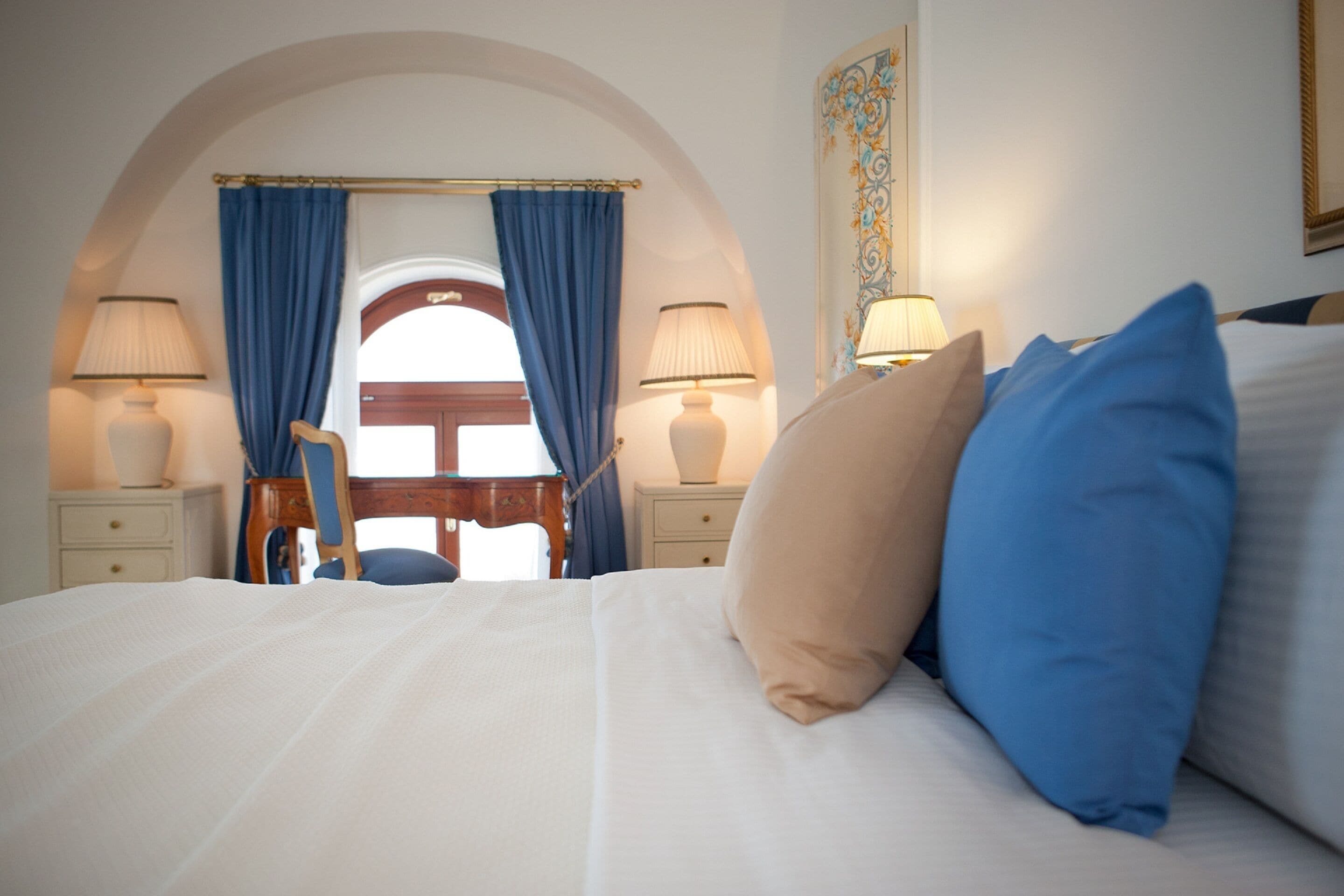 Une chambre d’hôtel avec un grand lit, deux lampes de chevet, une chaise en bois et une vue sur l’extérieur, visible à travers une porte en arc de cercle.