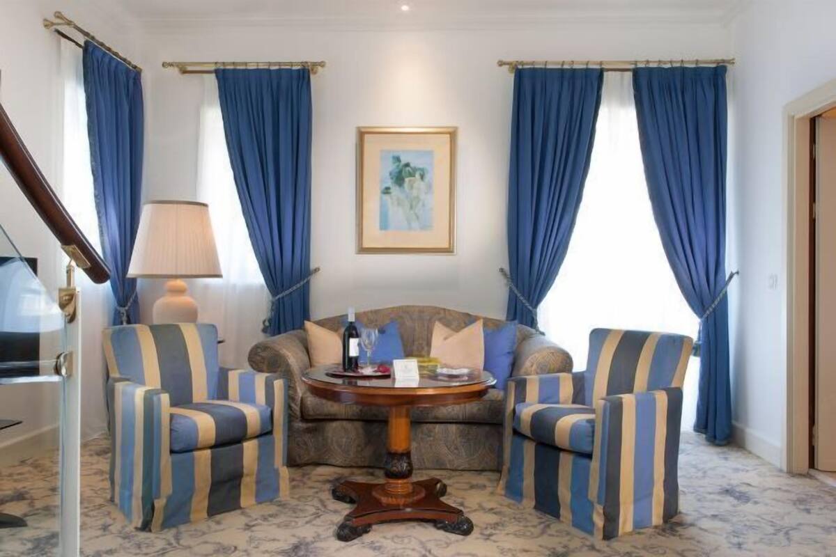 A room with a sofa, two striped armchairs, a round table, a lamp, and a painting on the wall.