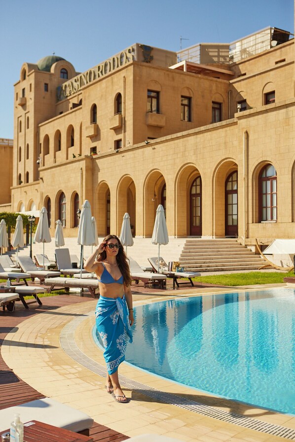 Un área de piscina con sillones reclinables y sombrillas, un edificio con ventanas arqueadas y un cartel que dice "CASINO RODOS".
