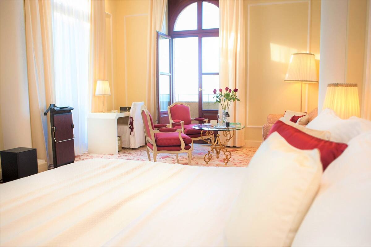 A hotel room with a large bed, a desk, a chair, a small table with flowers, and a lamp.
