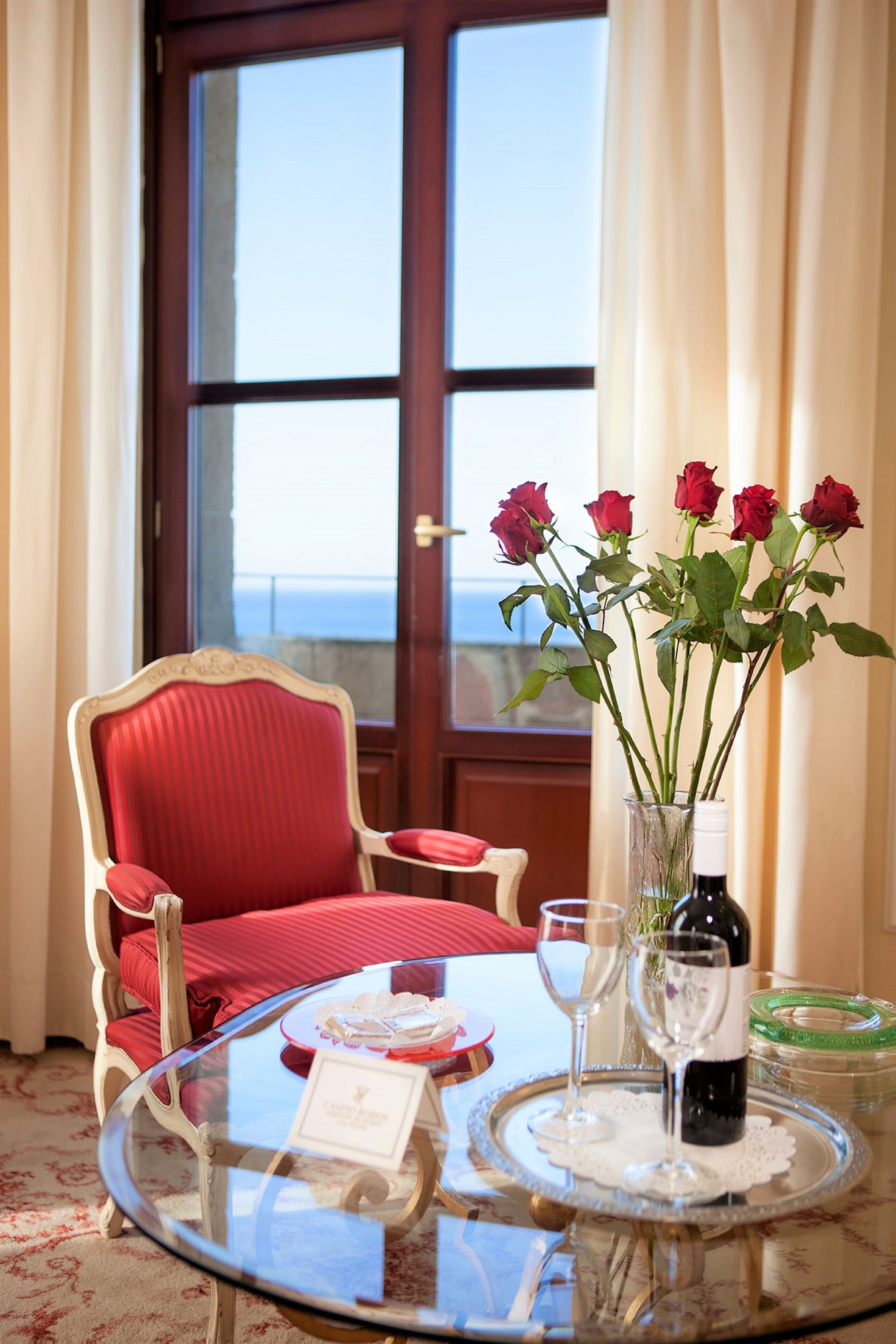 A dining area with a glass table, red upholstered chairs, a vase with red roses, a bottle of wine, and a view of the sea through a large window.