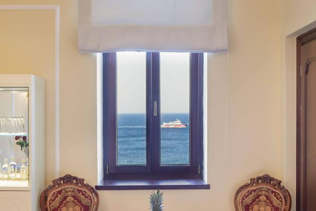 A room with a large window overlooking the sea, a shelf with bottles, and two ornate chairs.