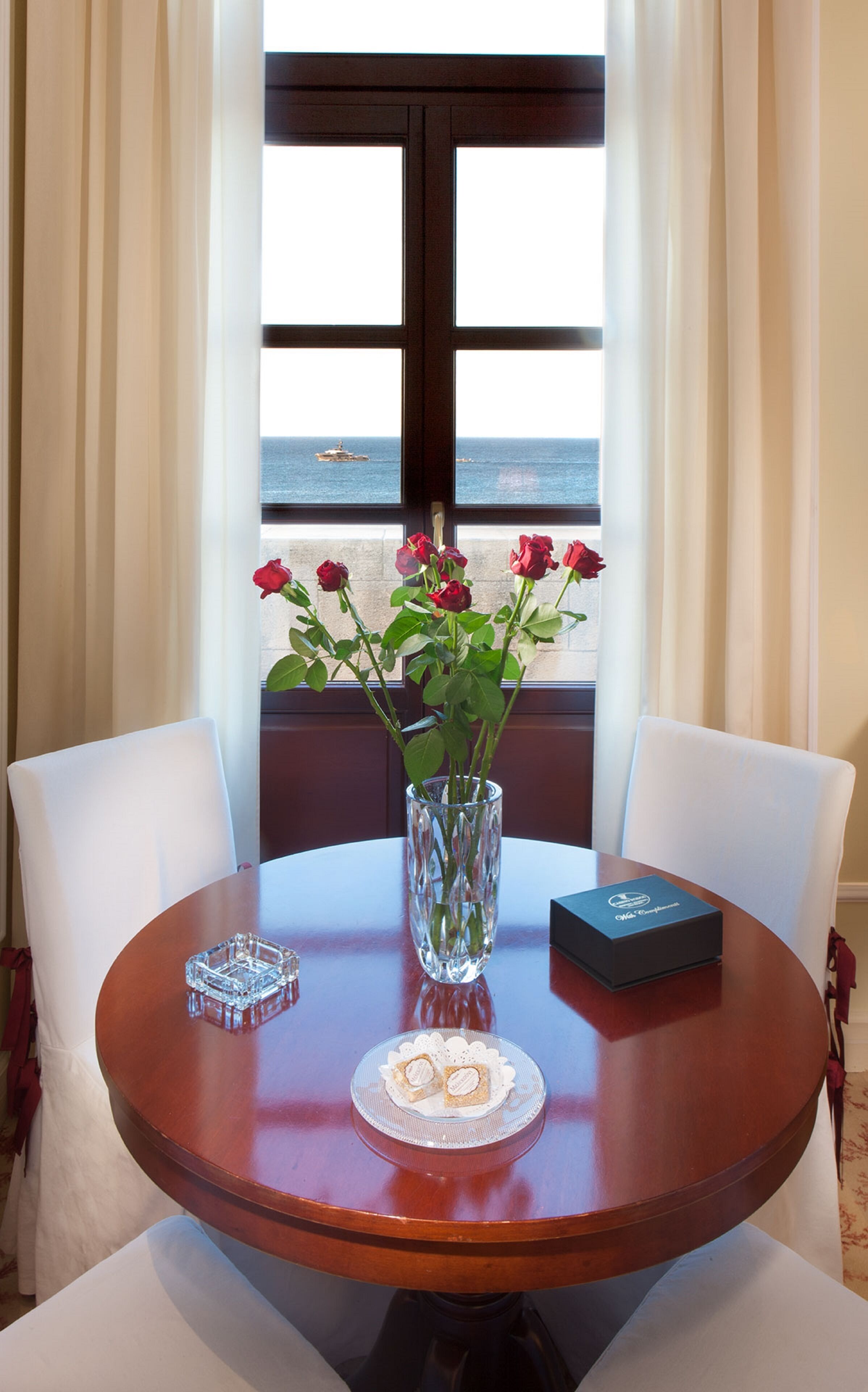 A round wooden table with a vase of red roses, a crystal ashtray, and a small box, set against a window with a view of the sea.