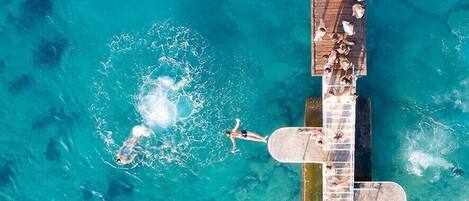 An aerial view of a clear blue pool with a wooden deck and a few people swimming and lounging.