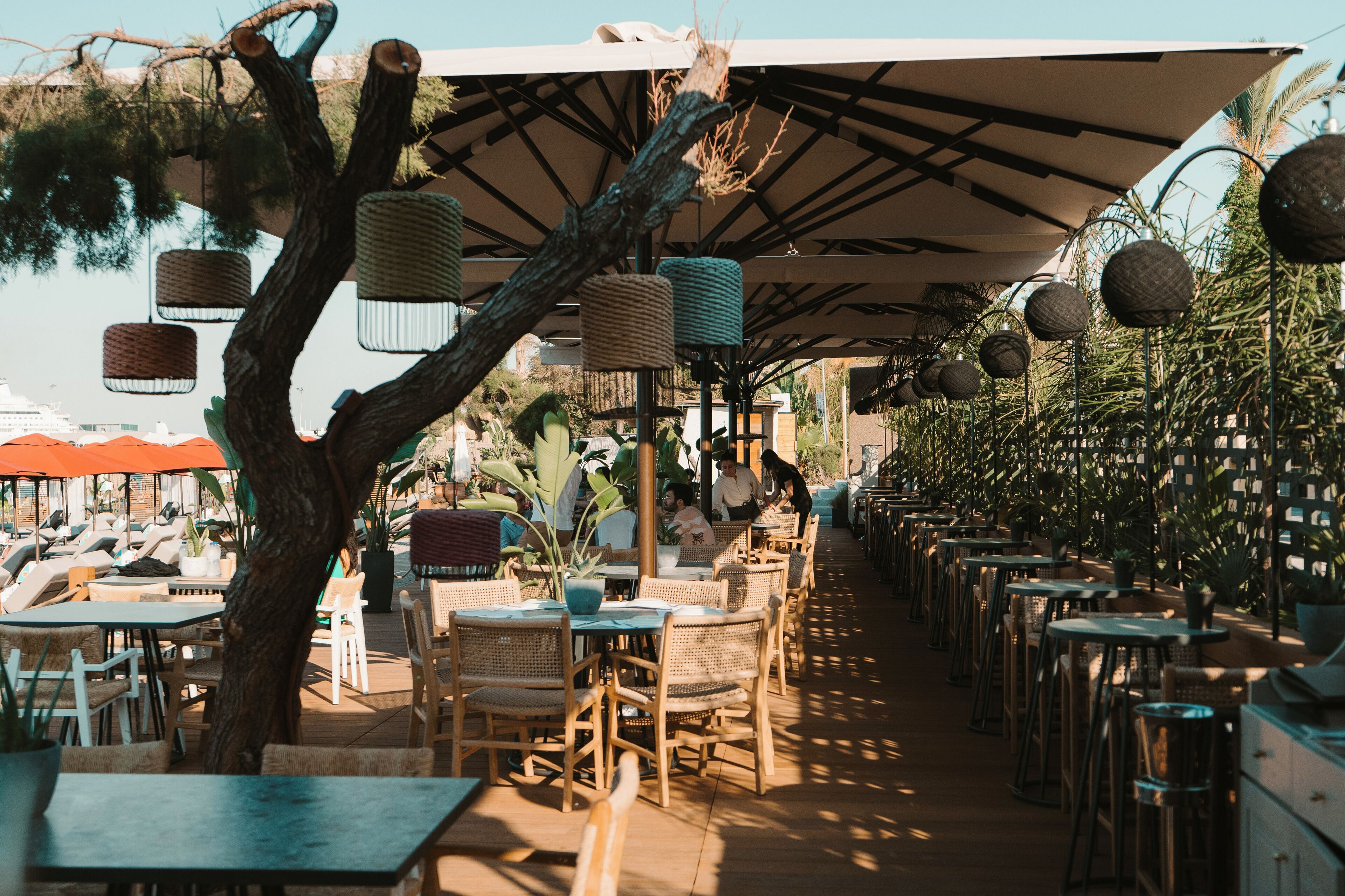 An outdoor café with wicker furniture, umbrellas, and hanging lanterns.