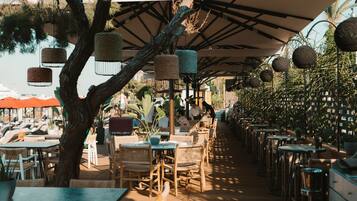 An outdoor café with wicker furniture, umbrellas, and hanging lanterns.