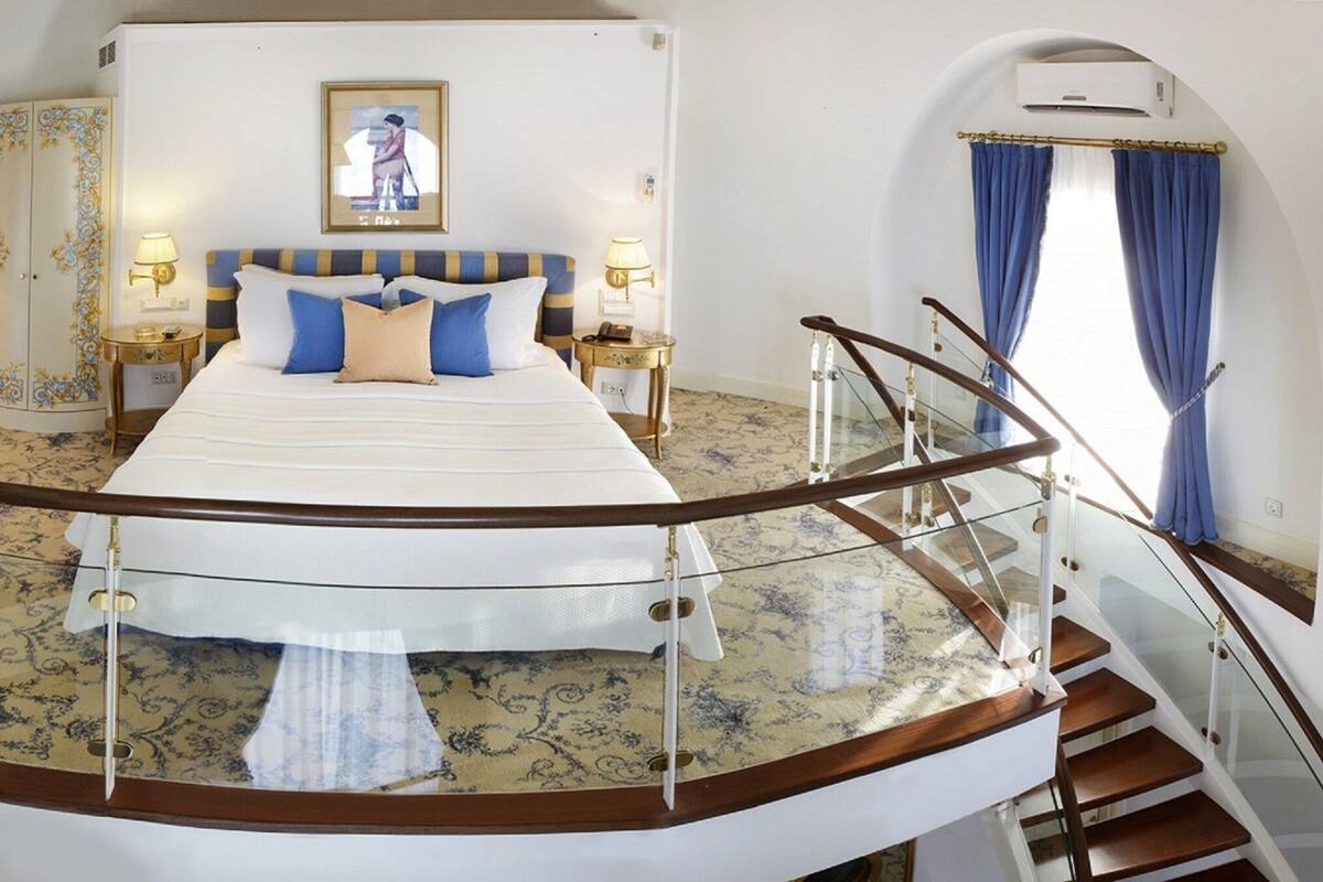 A spacious bedroom with a large bed, two bedside tables with lamps, a wardrobe, and a glass railing staircase.