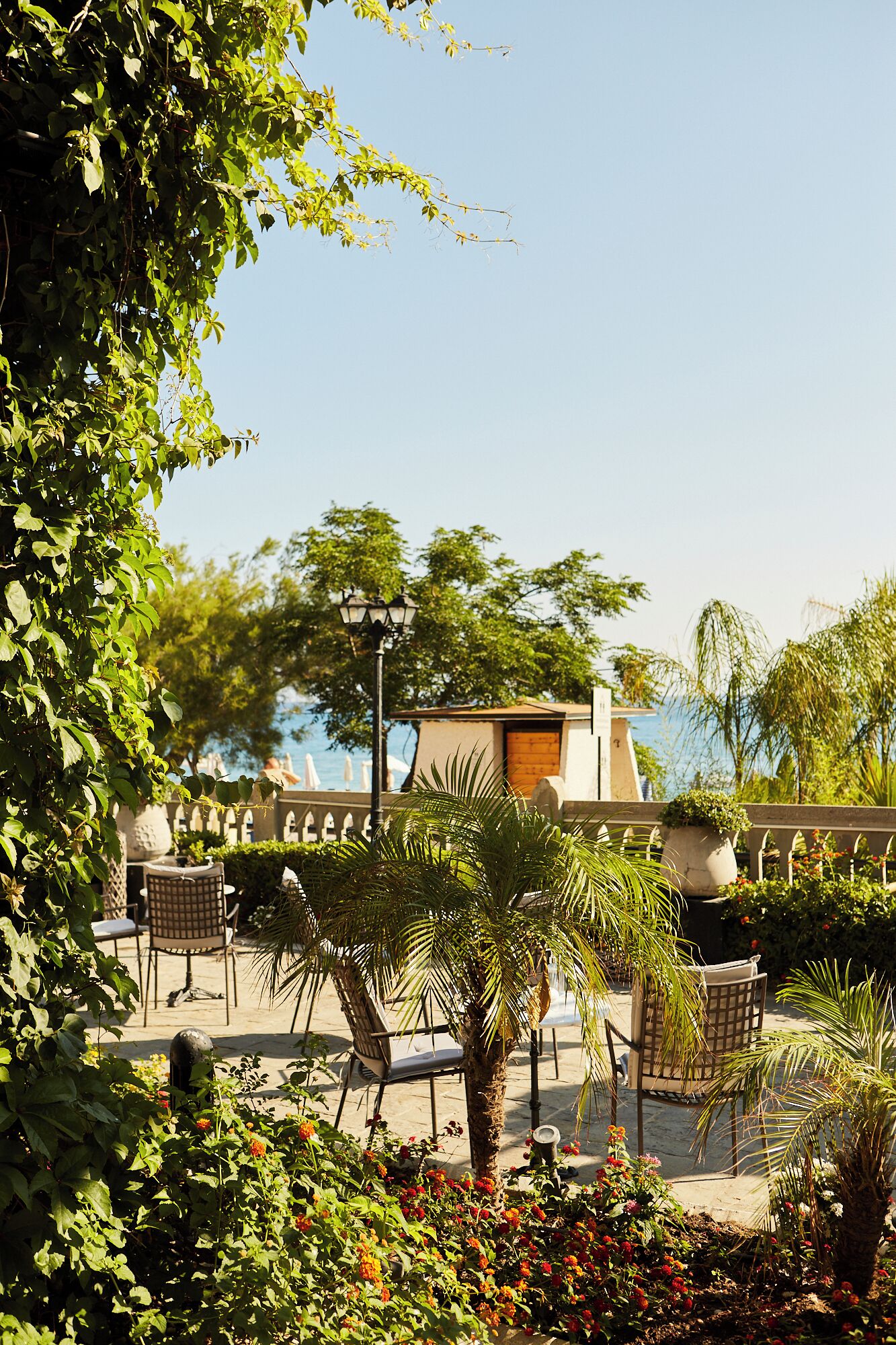 An outdoor seating area with chairs and tables, surrounded by lush greenery and a view of the sea.
