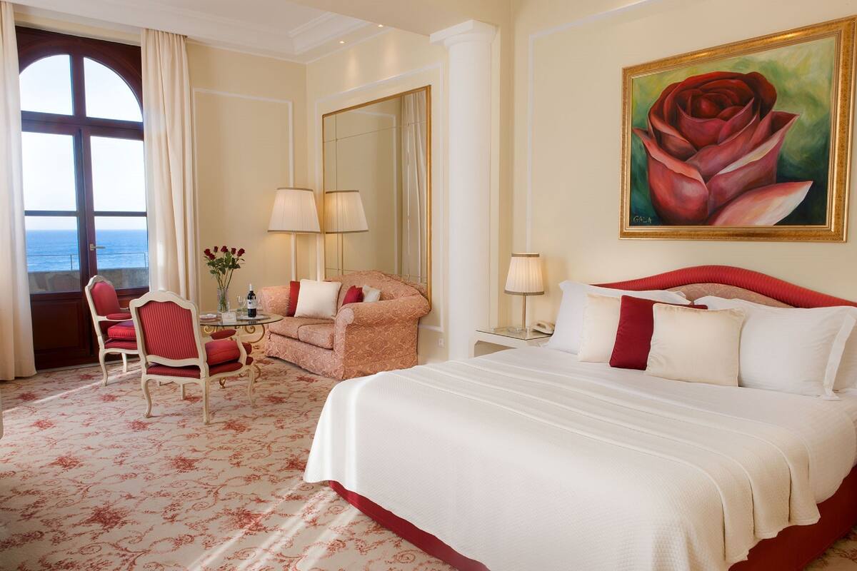 A hotel room with a large bed, a sitting area with a sofa and chairs, a painting of a rose, and a view of the sea through a window.
