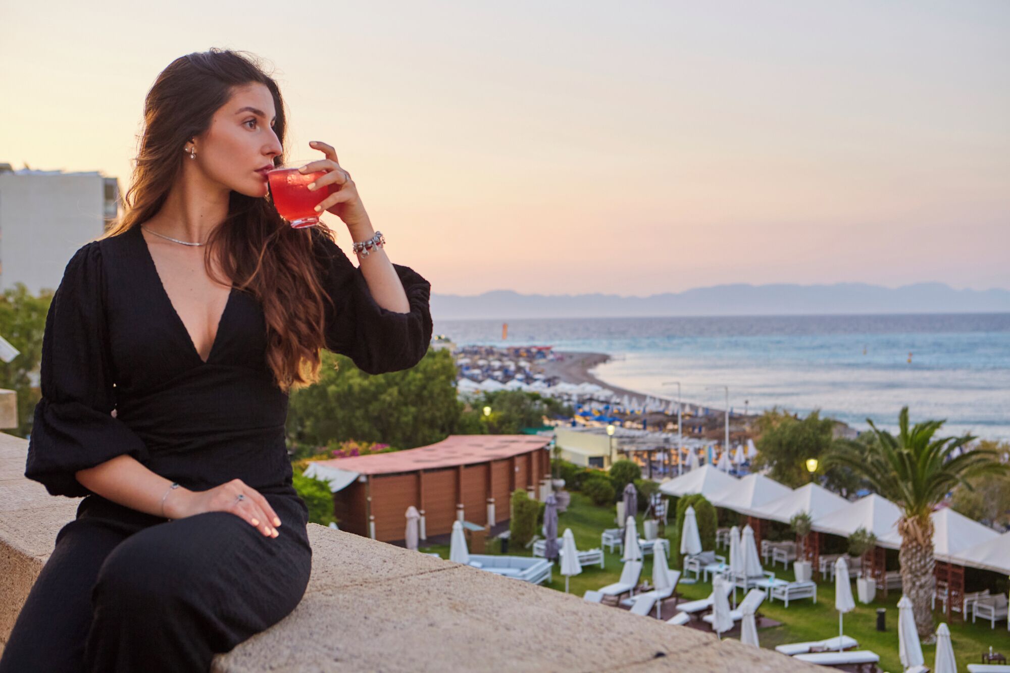 A person sitting on a stone ledge overlooking a beach with umbrellas and lounge chairs, suggesting a resort setting.