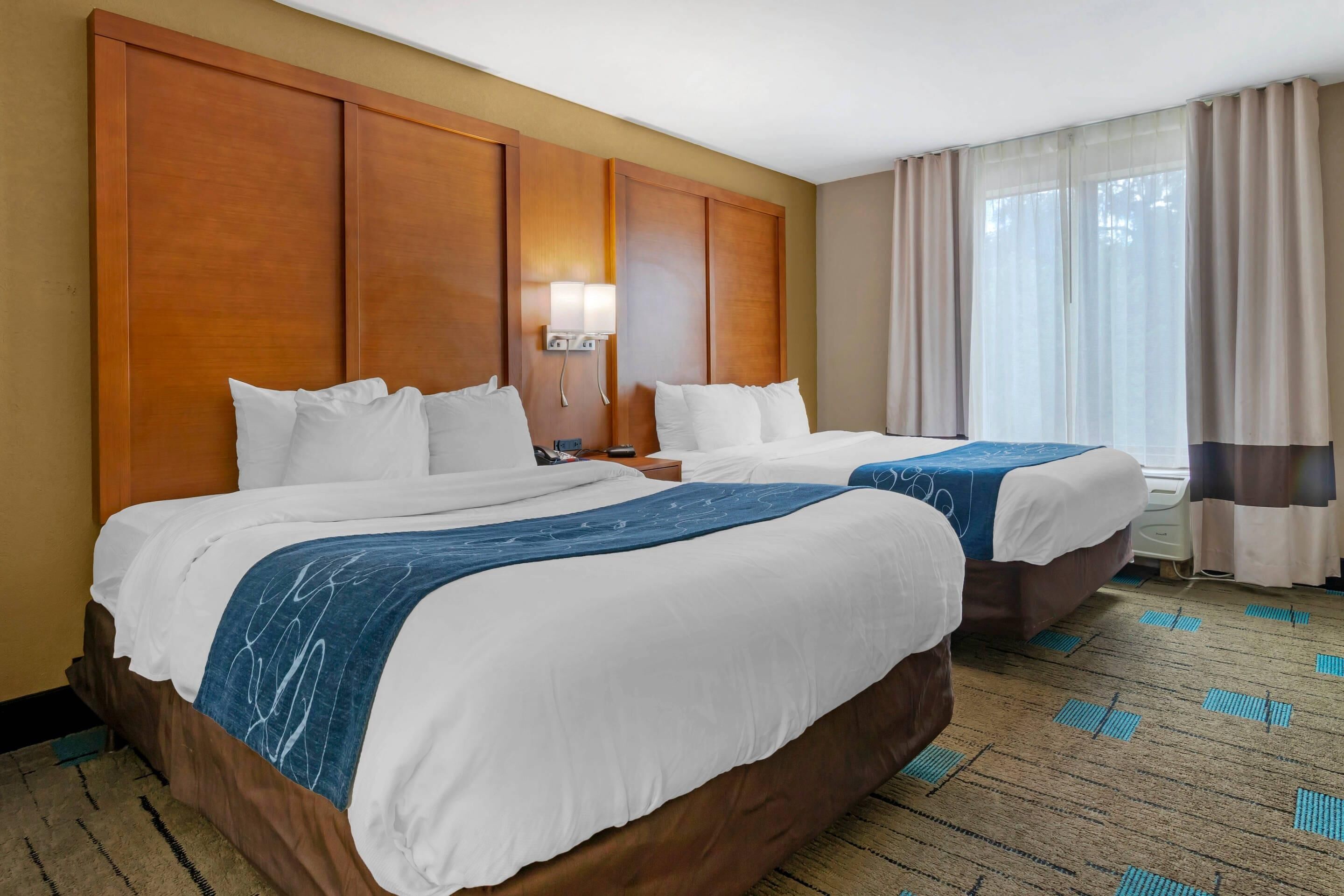 Habitación de hotel con dos camas, cabecera de madera, mesita de noche, lámpara y ventana con cortinas.