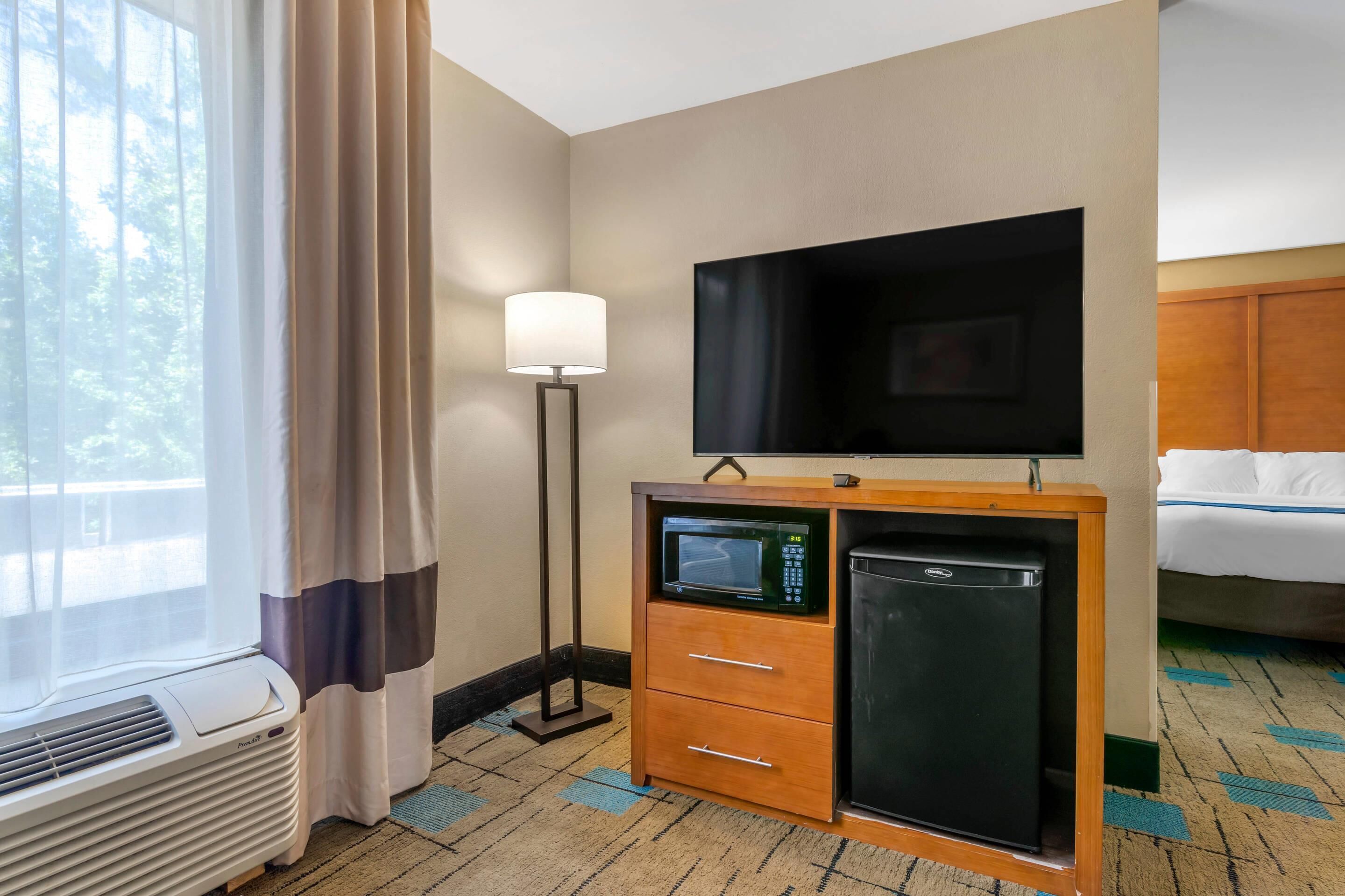 A hotel room with a flat-screen TV, a minibar, a heater, and a window with curtains.