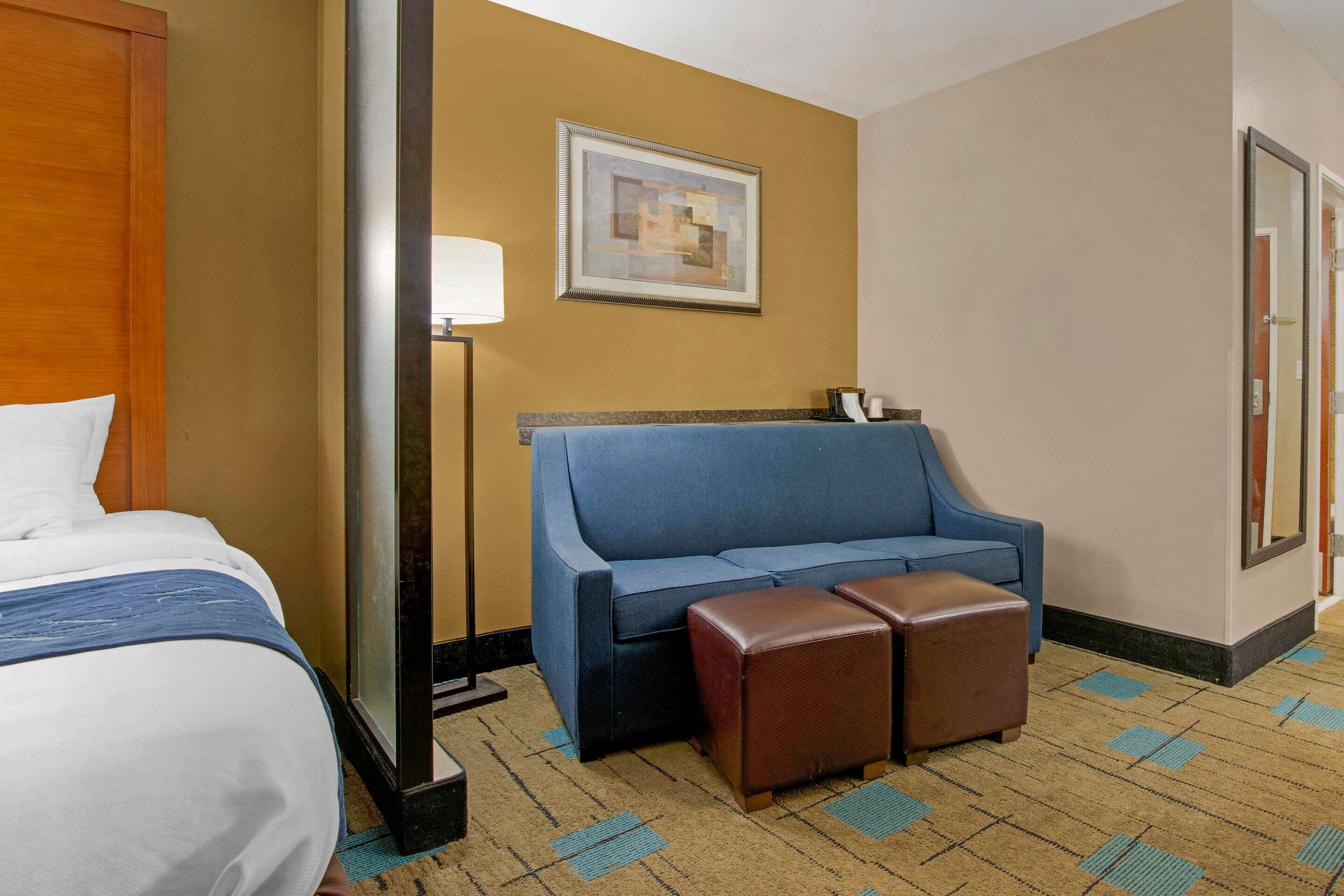Een hotelkamer met een bed, een blauwe bank, twee bruine poefs, een lamp en een lijst met een afbeelding aan de muur.