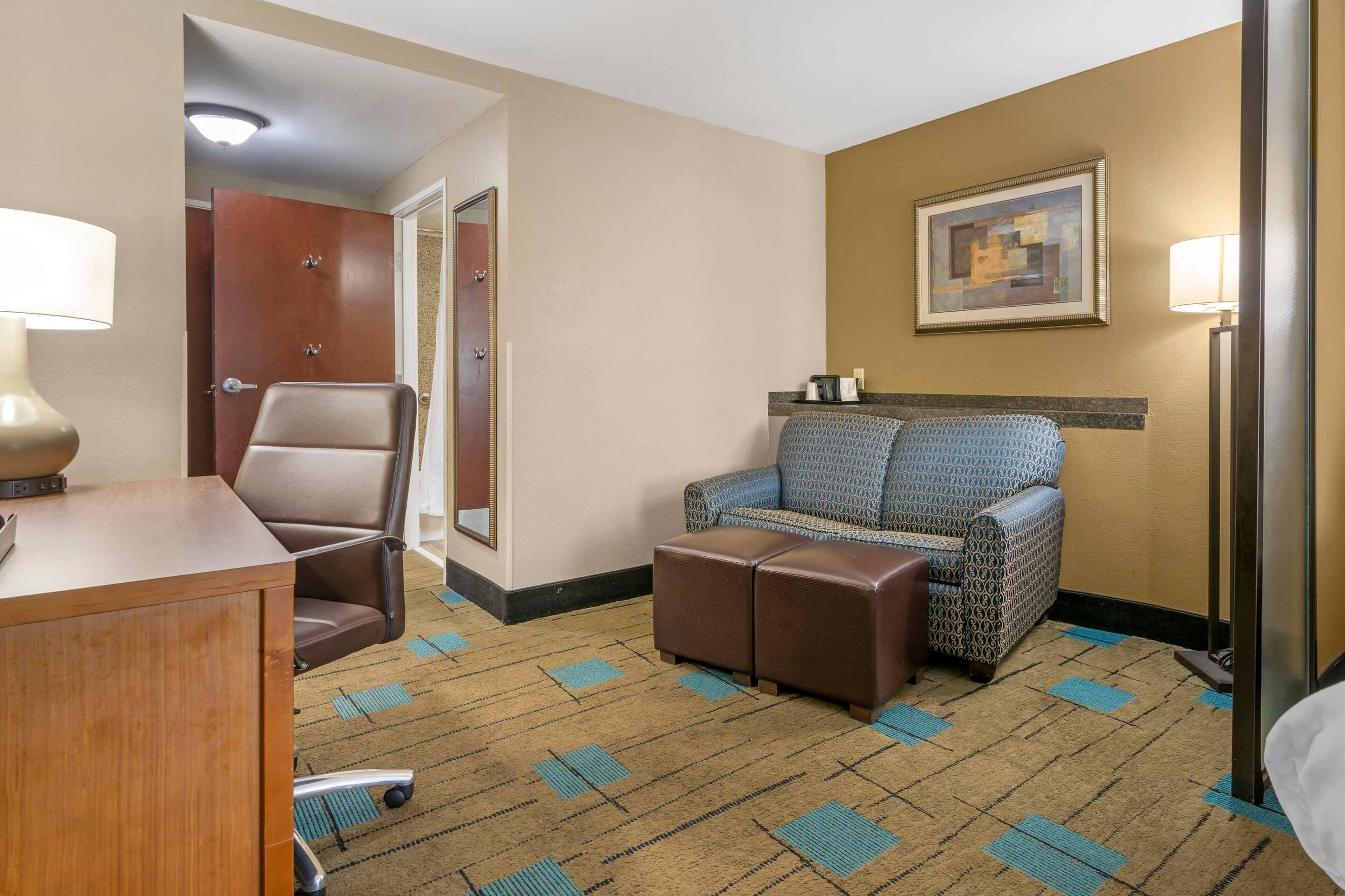 Habitación de hotel con escritorio, una silla, un sofá estampado, un otoman, un cuadro en la pared y una alfombra con diseño geométrico.