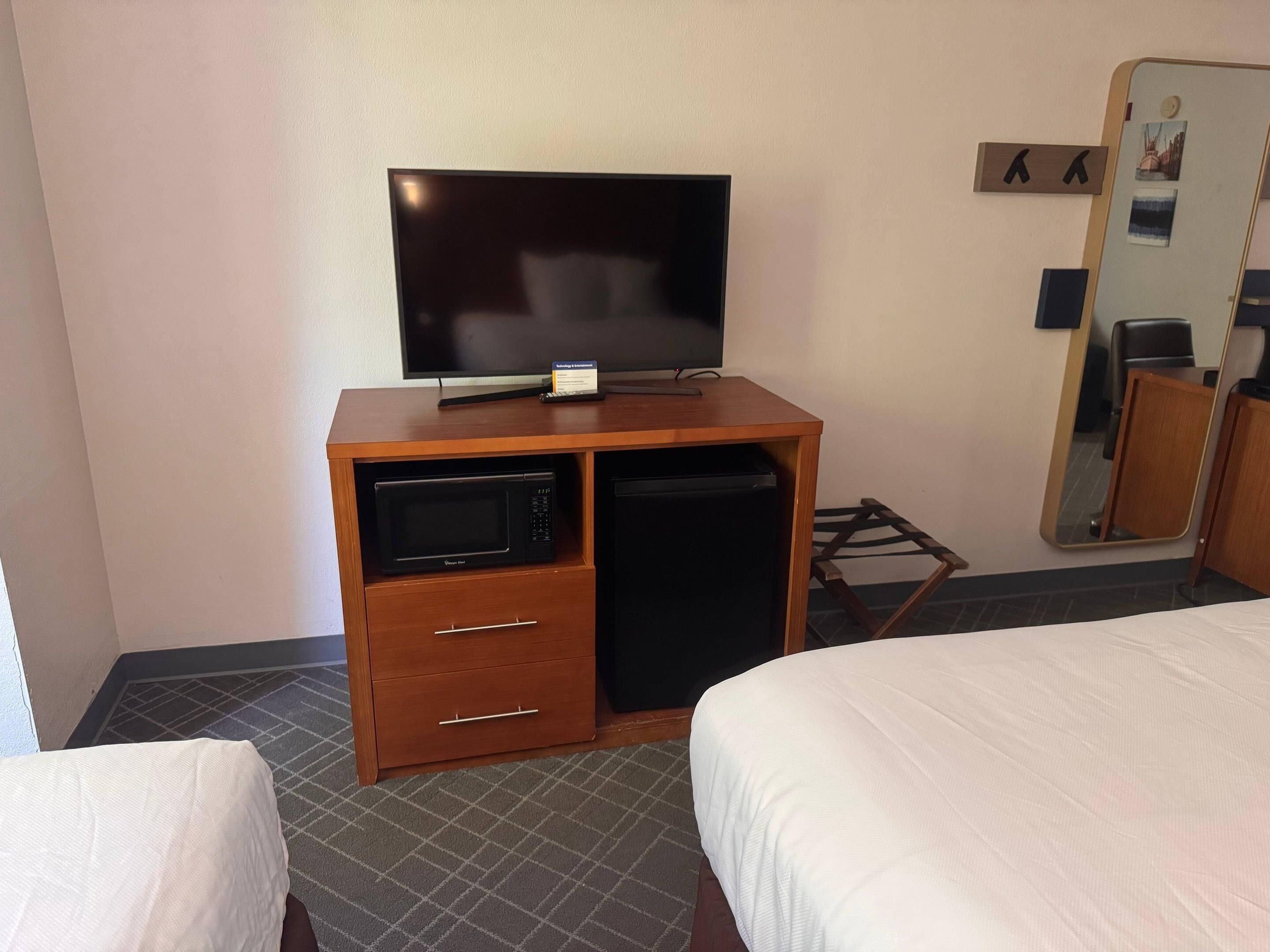 Ett hotellrum med en platt-TV placerad på ett träskåp, en mikrovågsugn, ett litet kylskåp, ett nattduksbord och en spegel.