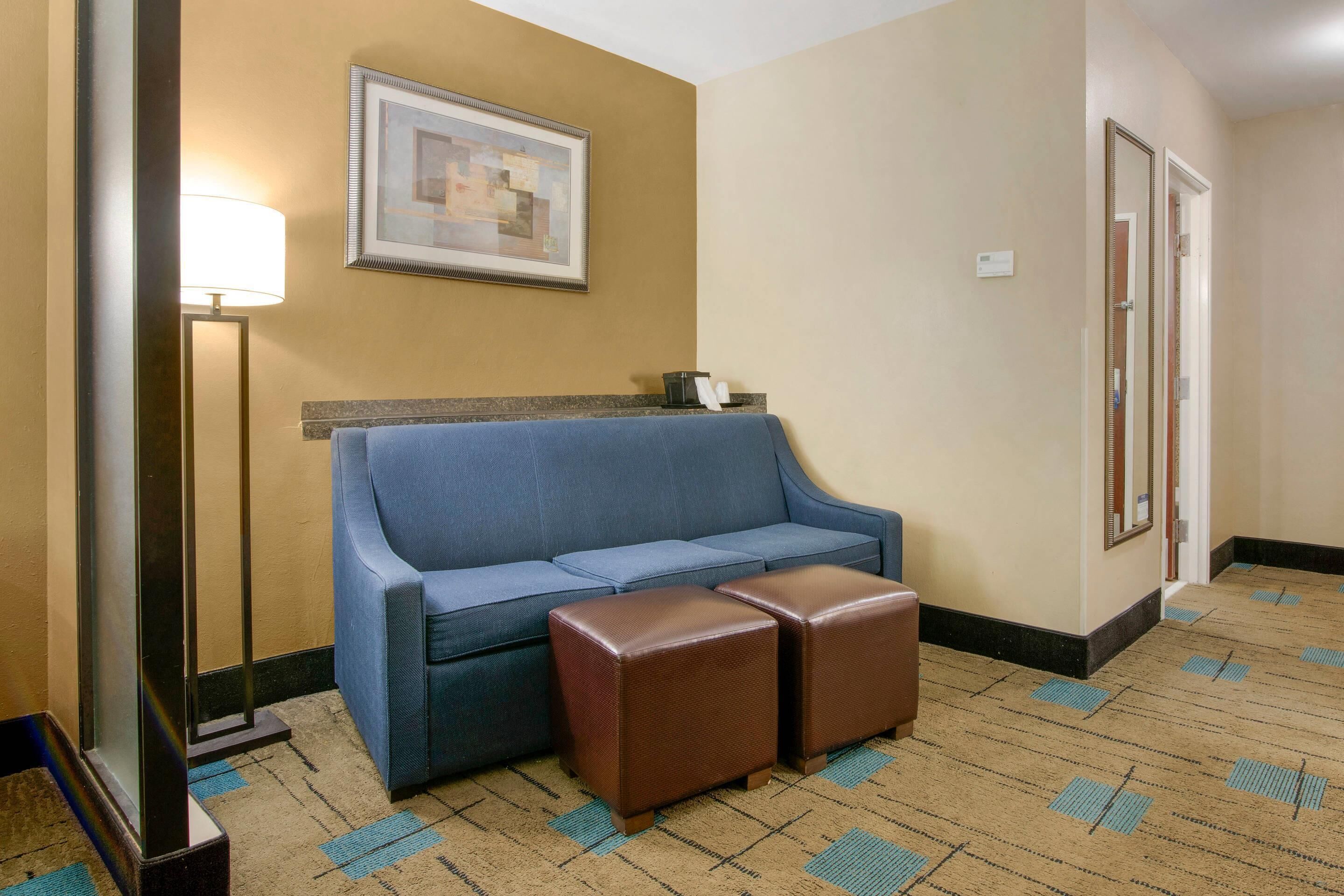 A hotel room with a blue sofa, a small brown ottoman, a framed picture on the wall, a floor lamp, and a door with a mirror.
