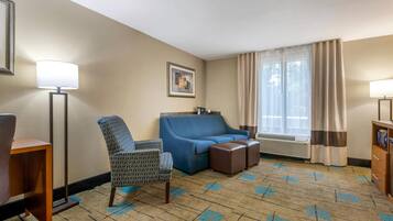 A hotel room with a sofa, armchair, and a desk with a lamp.