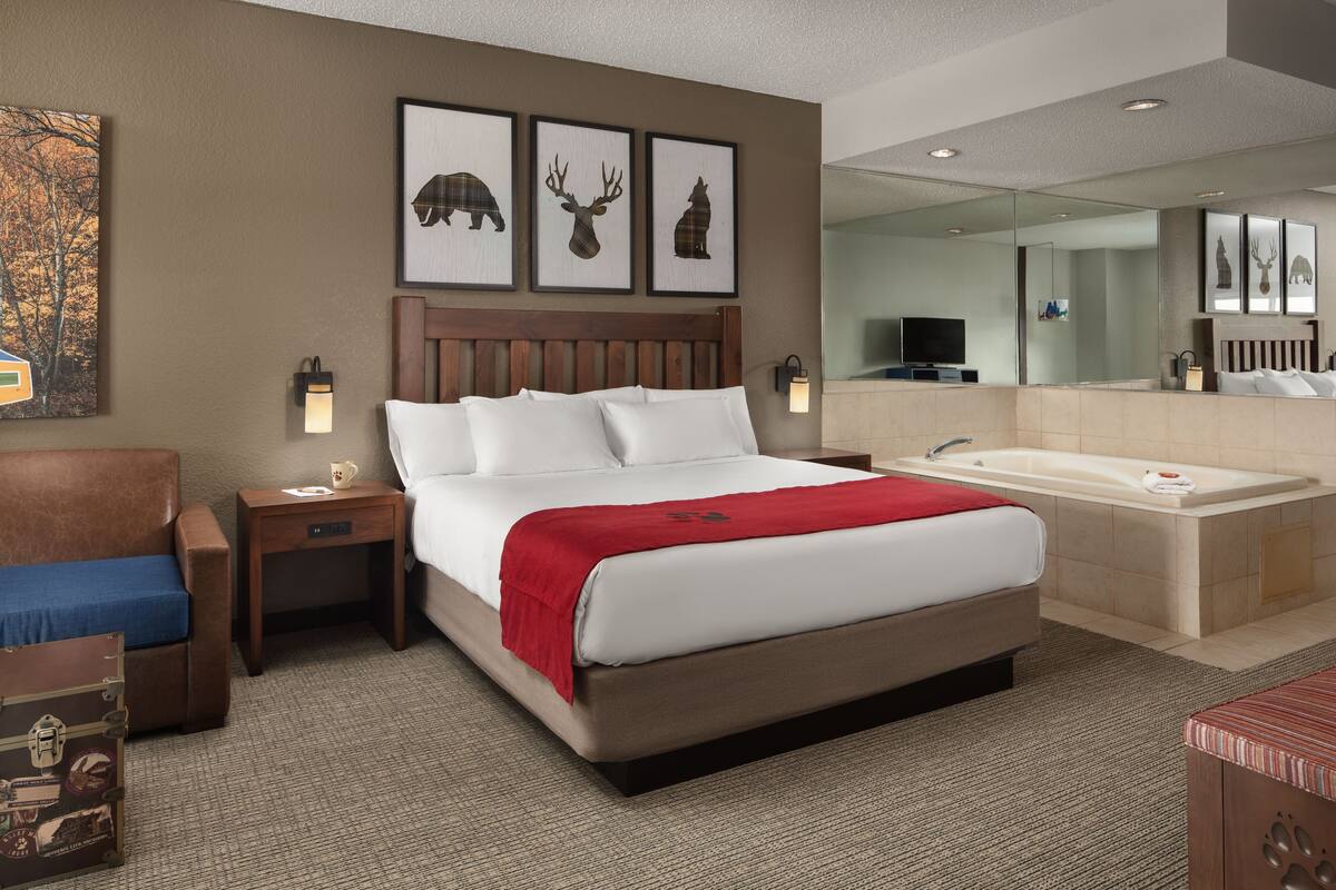 Ein Hotelzimmer mit einem großen Bett, einem Holzkopfteil, einer roten Tagesdecke, einem Nachttisch, einer Lampe, einem Koffer, einem Spiegel und einer Badewanne.