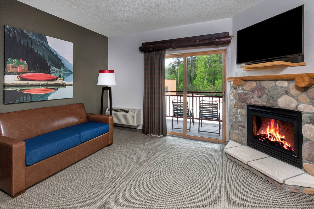 Ein gemütliches Zimmer mit Kamin aus Stein, einer Couch mit blauen Kissen, einem flachbildschirm-Fernseher über dem Kamin und einem großen Fenster mit Blick auf Bäume.