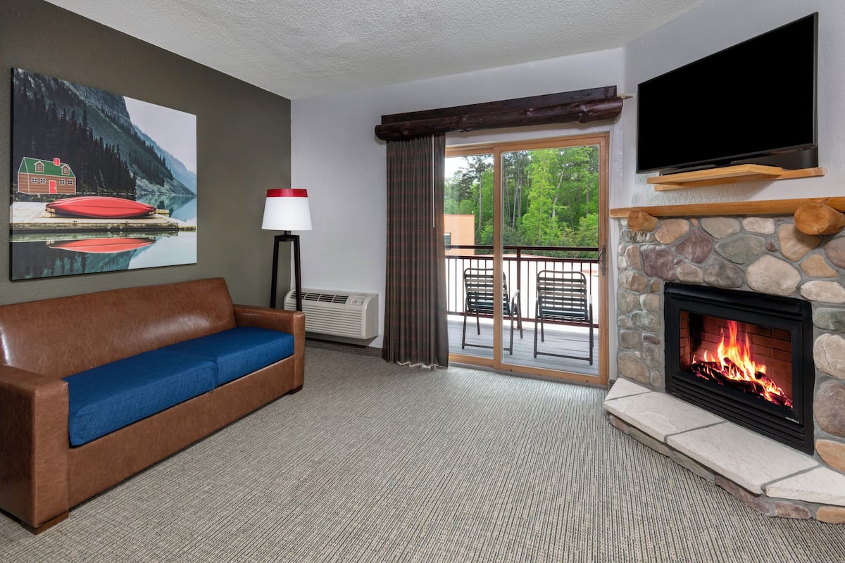 A cozy room with a stone fireplace, a sofa with blue cushions, a flat-screen TV mounted above the fireplace, and a large window with a view of trees.