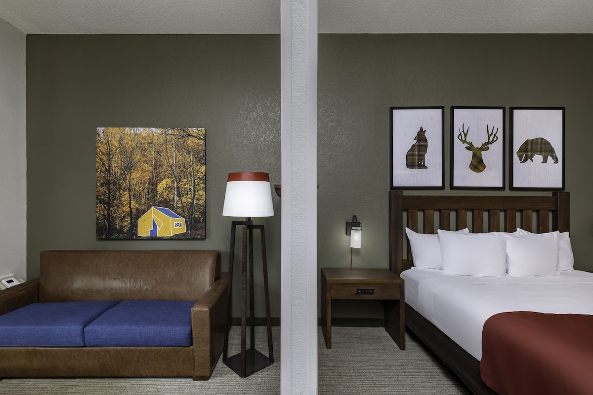 Ein Hotelzimmer mit braunem Sofa, einer Lampe, einem Nachttisch ebenfalls mit Lampe, einem Bett mit weißer Bettwäsche und drei gerahmten Bildern an der Wand.
