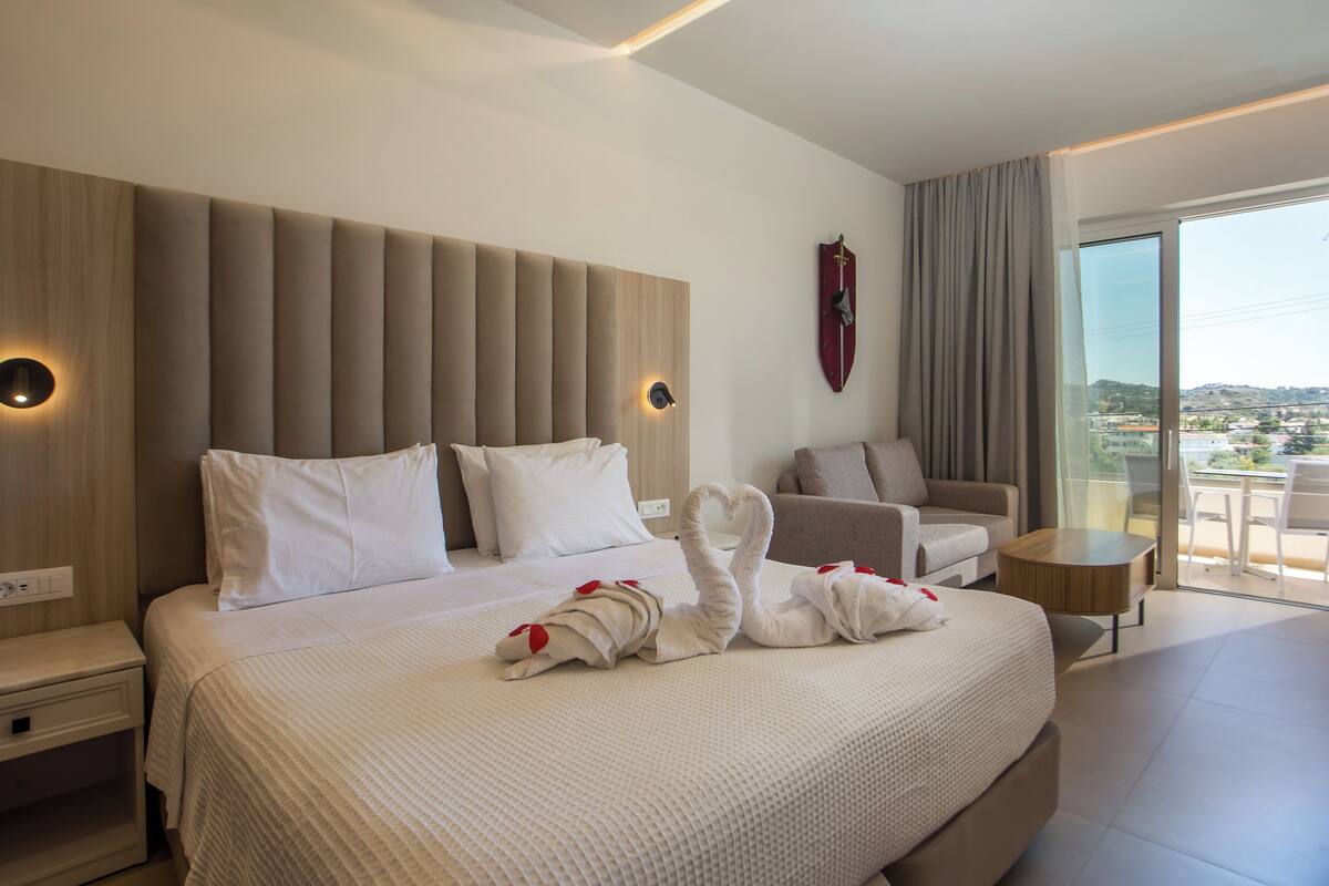 A hotel room with a large bed, a sofa, a small table, and a view of the city.