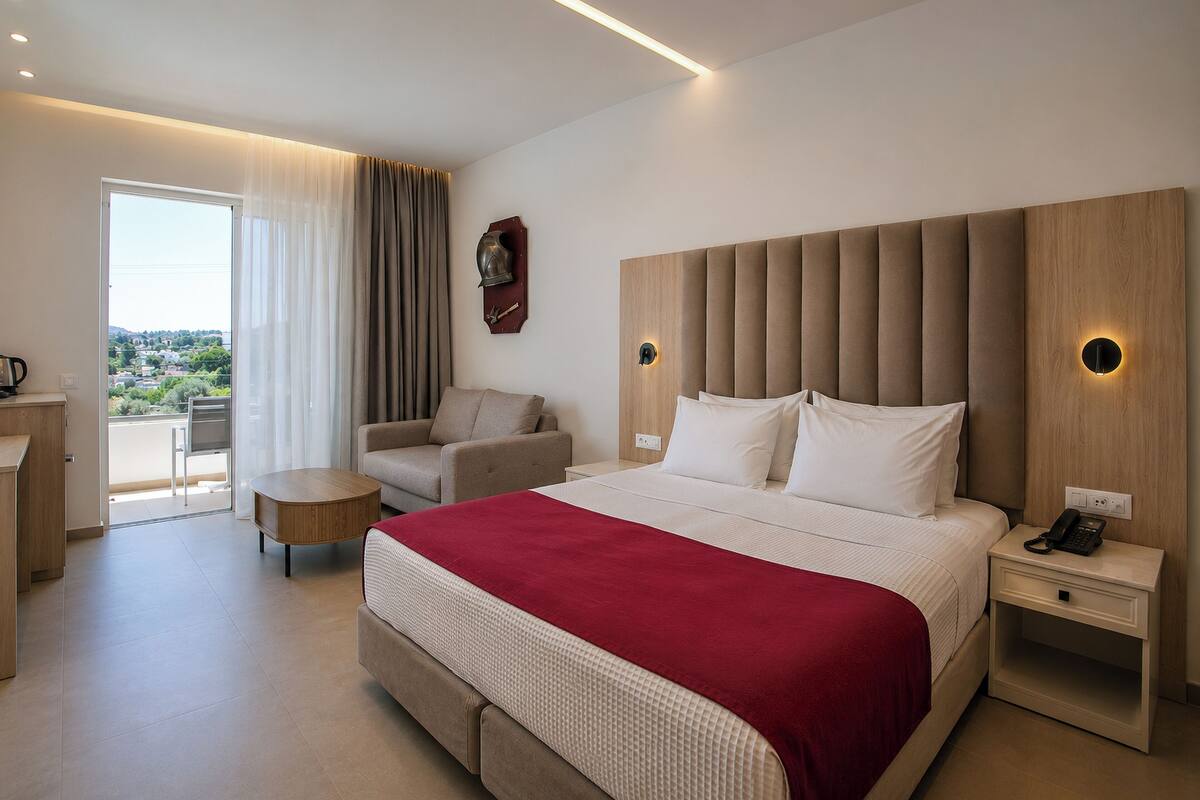 A hotel room with a large bed, a sofa, a small table, a desk, and a balcony with a view.