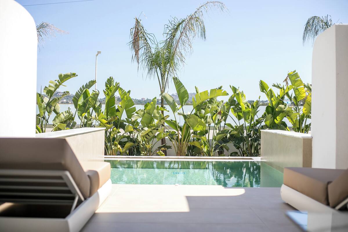 A poolside area with a view of a body of water, surrounded by lush greenery and modern architecture.