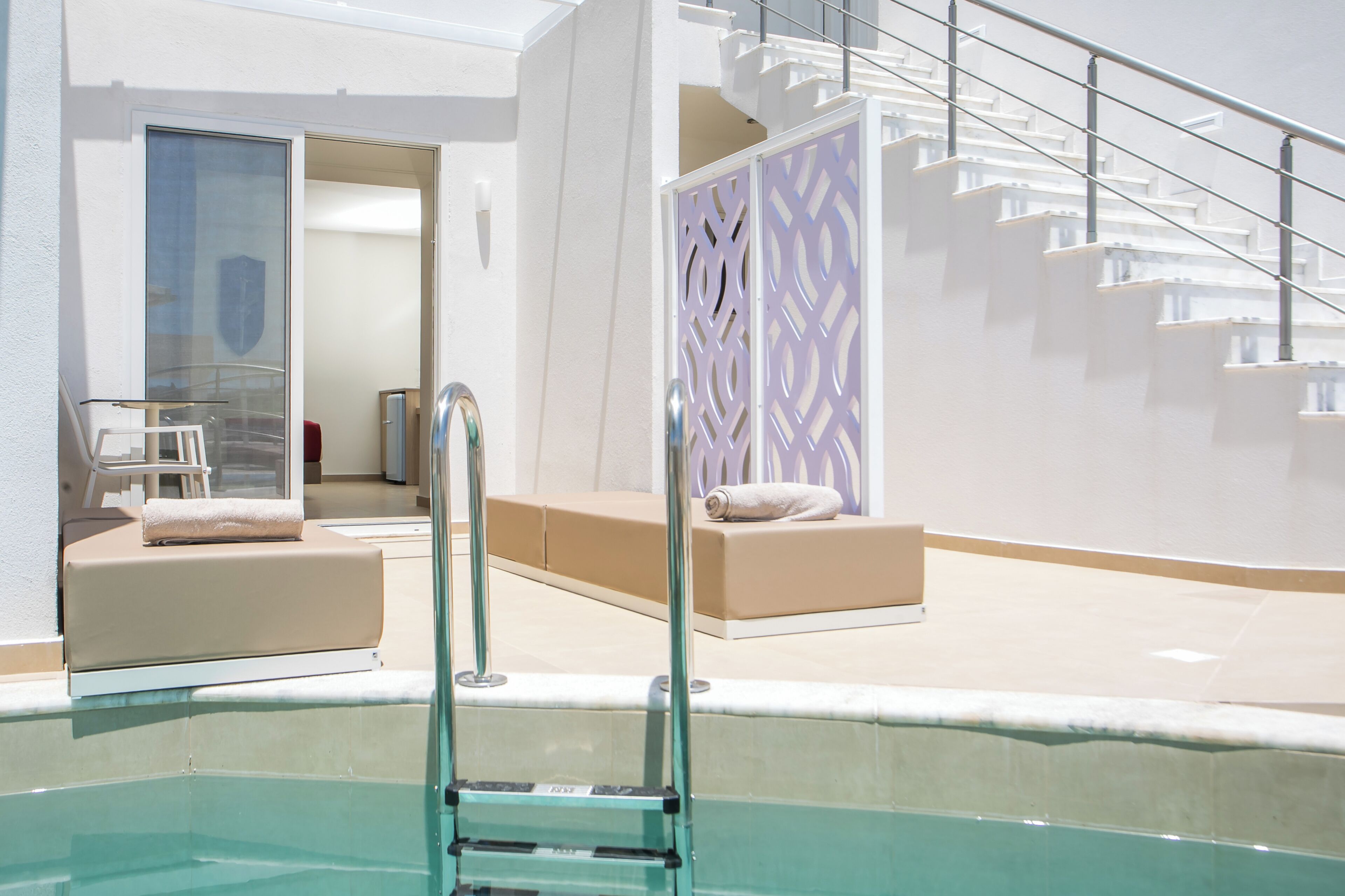 Un área de piscina interior moderna con barrera de cristal, una escalera y un espacio de descanso con cojines.