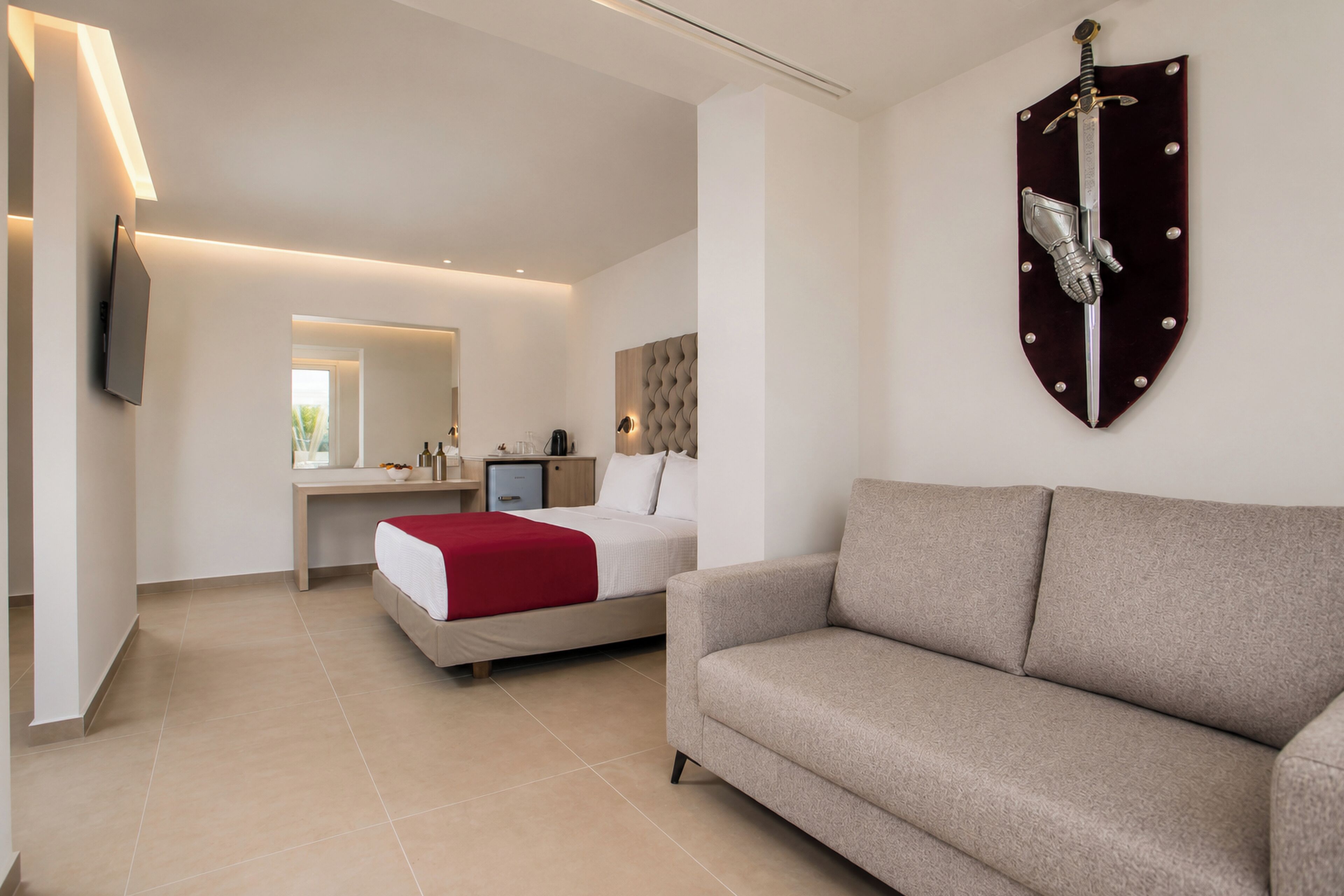 A modern hotel room with a bed, a sofa, a mounted sword, a flat-screen TV, and a mirror.