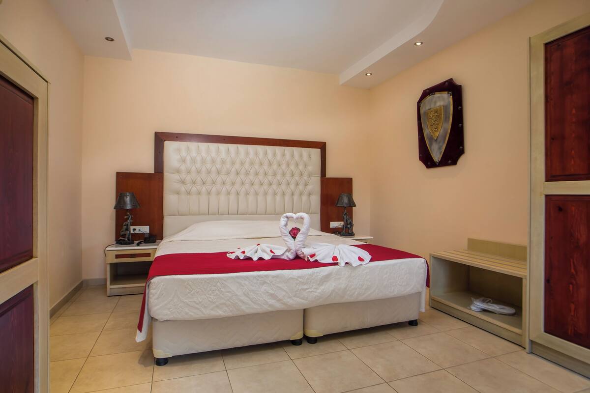 A hotel room with a large bed, two bedside tables, a wooden headboard, and a wall-mounted shield emblem.