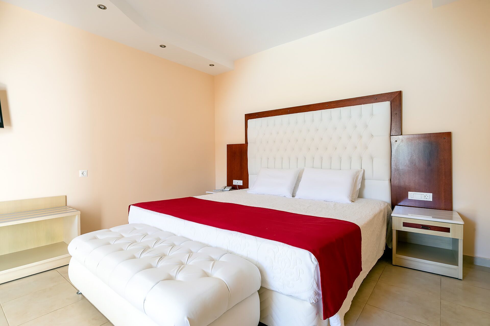 Habitación de hotel con una cama grande, una manta roja, un banco tapizado blanco, una mesita de noche de madera y una mesilla.