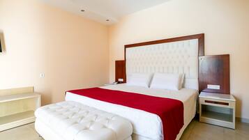 Habitación de hotel con una cama grande, una manta roja, un banco tapizado blanco, una mesita de noche de madera y una mesilla.