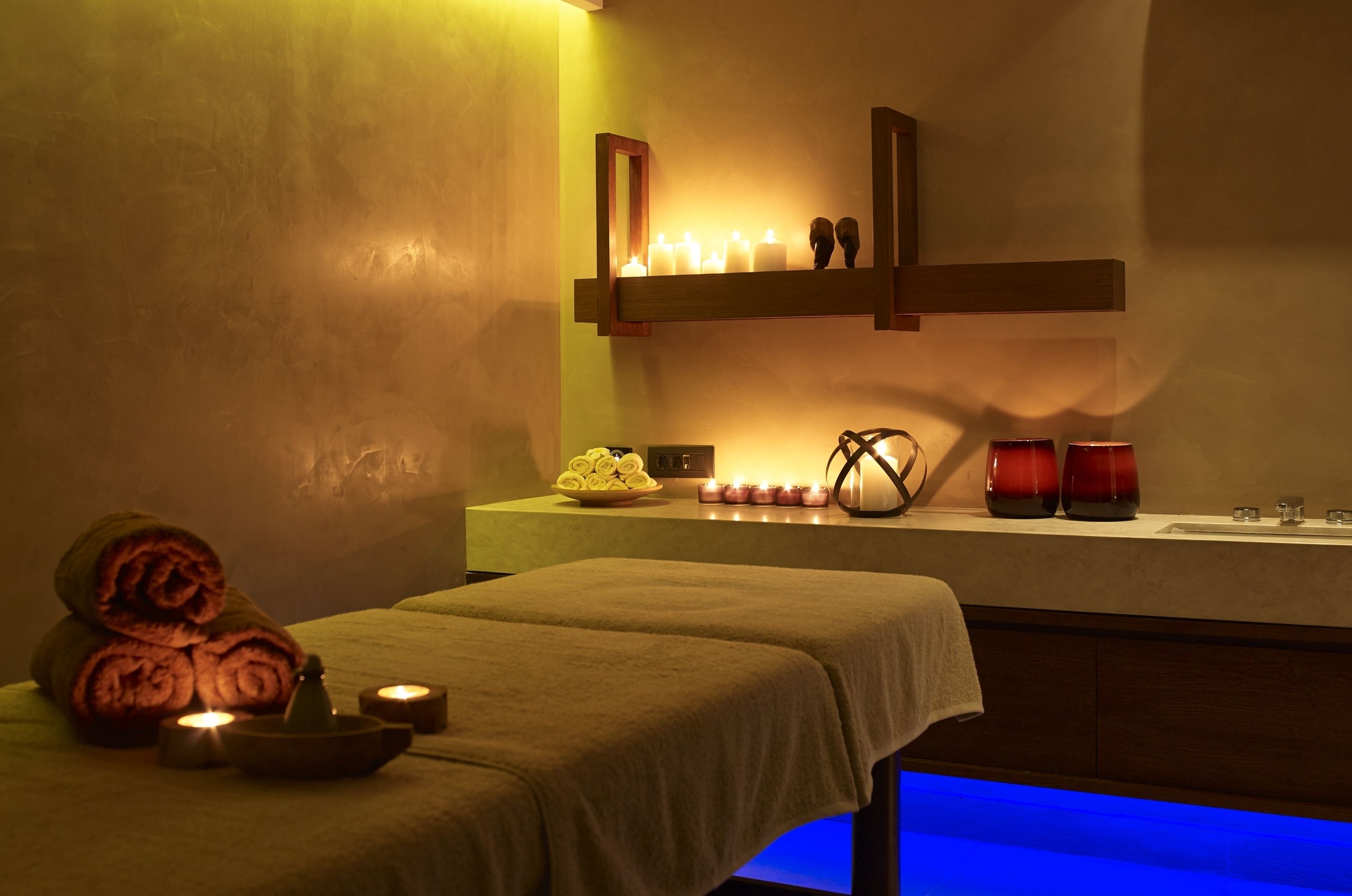Een spa-ruimte met twee massagetafels, kaarsen, een schaal gevulde handdoeken en een plank met kaarsen en een spiegel.
