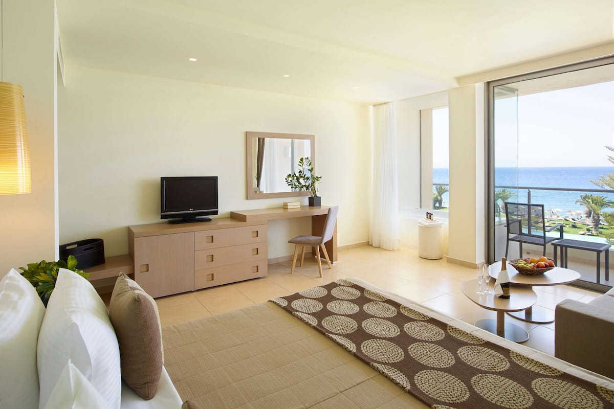 Habitación de hotel moderna con televisión de pantalla plana, un escritorio, una silla, un sofá, una mesa de comedor con un cuenco de fruta y vista al océano.