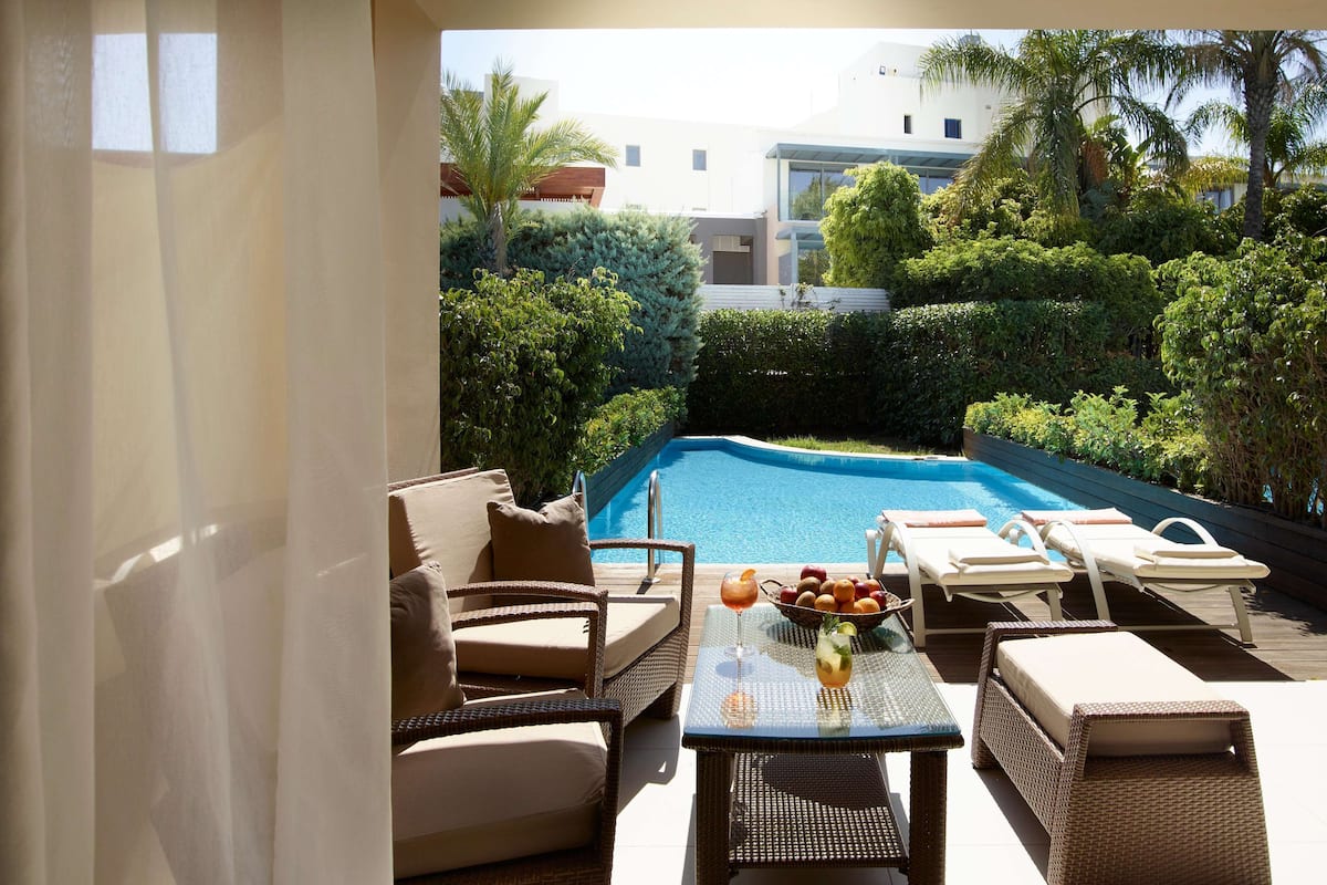 Zona junto a la piscina con mobiliario de mimbre, una mesa con frutas y bebidas, y vistas a una piscina rodeada de vegetación.